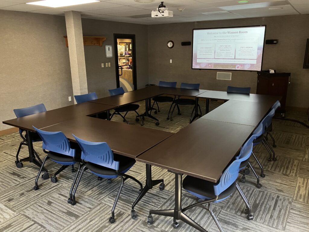 Empty board room with 8 roll-away tables and seats for up to 25, image projected on screen reads: Welcome to the Wasson Room!