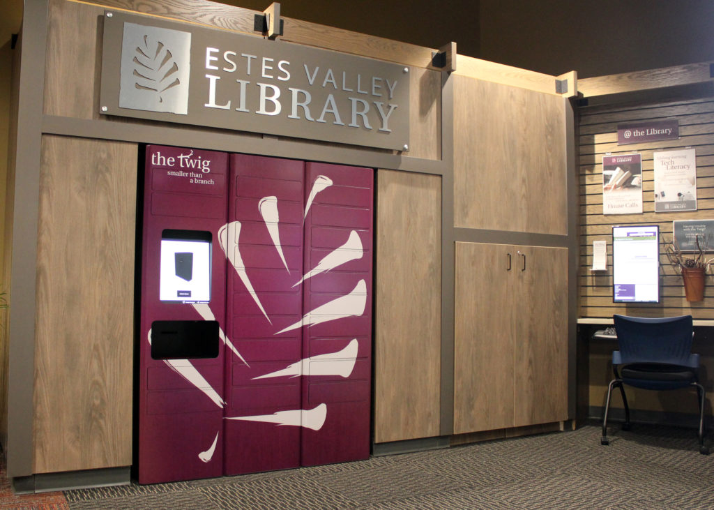 A wooden cabinet with sign and logo, Estes Valley Library, surrounds a purple book locker with the white Estes Valley pine cone logo and a computer screen