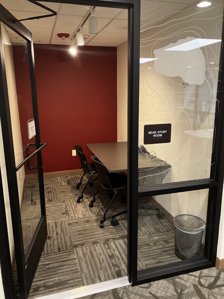 An open door welcoming to a study room with one table, two chairs, and a port for charging devices.