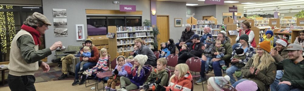 A man reads aloud from the children's book The Polar Express to an audience of children and adults dressed in festive holiday attire.