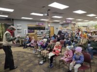 A man reads aloud from the children's book The Polar Express to an audience of children and adults dressed in festive holiday attire.